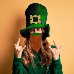 Beautiful brunette woman wearing green hat with clover celebrating saint patricks day shouting with crazy expression doing rock symbol with hands up. Music star. Heavy concept.